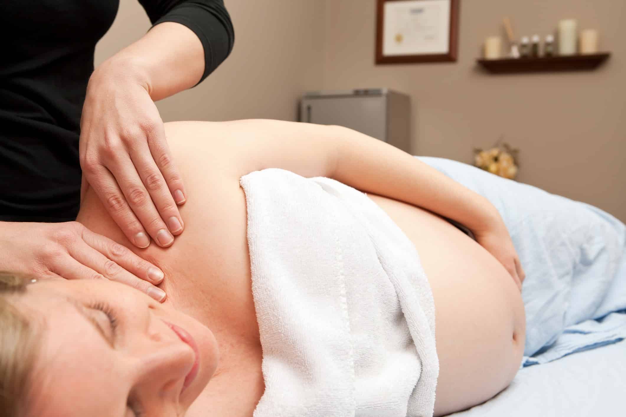 A pregnant woman is receiving a prenatal massage from a female massage therapist. This type of massage is used to relieve tension, stress and discomfort during pregnancy. The background includes a stone/towel warmer on the left, flowers, a blurred accreditation document and a shelf with candles and oils.